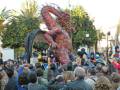 Una figura gigante de un dragón rojo con ojos brillantes y una boca abierta mostrando dientes esculpidos, rodeada por un grupo de personas en una celebración festiva. El ambiente está lleno de color y diversión, con árboles frutales en el fondo.