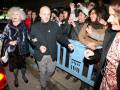 Una anciana con abrigo de piel y vestido negro, un hombre con chaqueta negra y pantalones grises caminando entre una multitud. La imagen muestra un ambiente social, posiblemente en una celebración o evento público.