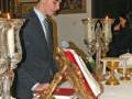 Un joven en traje negro está leyendo un libro abierto sobre una mesa de madera con ramas. A su lado, hay candelabros dorados y un cuadro en la pared. La escena parece ser en una habitación elegante, posiblemente un salón o biblioteca.