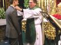 Un sacerdote bendice a un hombre en una ceremonia religiosa, rodeados por adornos dorados y flores rojas.