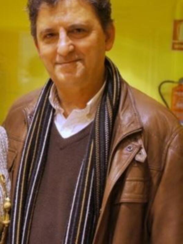 Un hombre con una chaqueta de cuero y una camisa de vestir, posando frente a una pared amarilla.