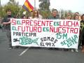 Una multitud de personas sostiene un gran cartel que dice "Sí, el presente es lucha. El futuro es nuestro. Participa. Somos el 99%. Sevilla Este". La imagen muestra a manifestantes en una calle, con banderas y pancartas.