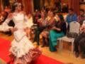 Una mujer en un vestido de flamenca blanco y rosa, con una corona de flores, baila en un escenario rojo. Los asistentes observan la actuación con interés. La imagen transmite un ambiente festivo y cultural, probablemente en una fiesta o evento de baile.