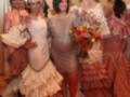 Una mujer en un vestido de lentejuelas posa junto a una mujer con un vestido blanco y otro vestido de color naranja. Todos están en un escenario con flores y decoraciones.