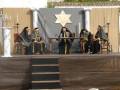 Una escena ceremonial con cuatro figuras vestidas en trajes tradicionales, sentadas frente a una bandera estrella de David. En el fondo, se observan cortinas y un altar con velas.