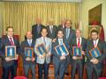 Un grupo de personas en trajes posa para una foto. Algunos llevan medallas con estatuillas y otros tienen insignias en sus manos. La imagen se toma en un entorno formal, posiblemente una sala de conferencias o oficina. La decoración incluye cortinas doradas y un cuadro en la pared.