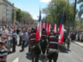 Una procesión militar con soldados montando caballos, banderas y un féretro. Un grupo de personas observa la procesión desde el lado del camino.