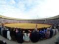 Una plaza de toros con una gran audiencia sentada en gradas, donde un torero se encuentra en el centro. La arena es de color amarillo y la gente está observando atentamente.