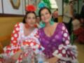 Dos mujeres en trajes típicos flamencos sonríen para la foto. La imagen está ambientada en un ambiente festivo con decoraciones y otros asistentes al fondo.