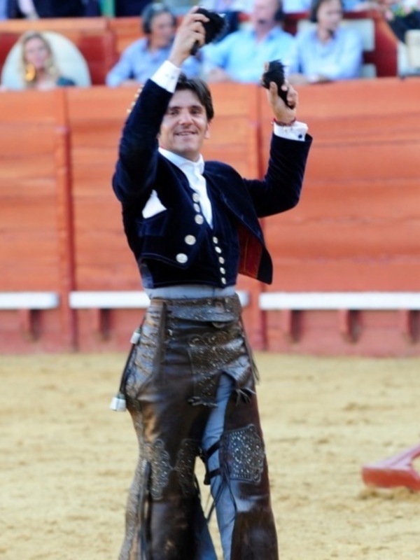 Un torero en una corrida de toros, con chaqueta azul y pantalones negros, levanta el puño en señal de victoria. La arena es de arena y las gradas son naranjas.