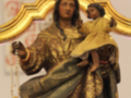 Una estatua de la Virgen María vestida en oro, sosteniendo a un niño Jesús. La estatua está coronada y lleva una corona dorada, con un fondo que parece ser de madera. La estatua está en una iglesia o lugar religioso, con un cartel de fondo que parece ser una inscripción en español.