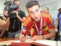 Un futbolista en camiseta de la selección española firma un documento en una mesa, mientras un cámara de televisión lo filma.
