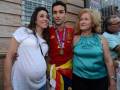 Un hombre vestido con una camiseta de fútbol roja y amarilla, posando junto a dos mujeres. La persona en el medio lleva una medalla dorada y sostiene un pequeño banderín español. La imagen está tomada en un entorno urbano con edificios de fondo.