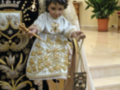 Un niño vestido con un traje tradicional, posiblemente para una celebración religiosa o festiva, está sosteniendo un cáliz en su mano derecha. El niño lleva una saya bordada con detalles dorados y un sombrero que parece ser parte de su vestimenta. A su lado, se puede ver una figura con un escudo y una corona, probablemente representando a un personaje importante o líder. La imagen se encuentra en un entorno interior, con una planta y escaleras al fondo.