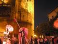 La Giralda, la catedral de Sevilla, iluminada en la noche. Una figura con una espada y otro personaje con un sombrero, posiblemente en una parodia del espectáculo de fuegos artificiales. La gente observa la escena en el fondo, con árboles y edificios antiguos.
