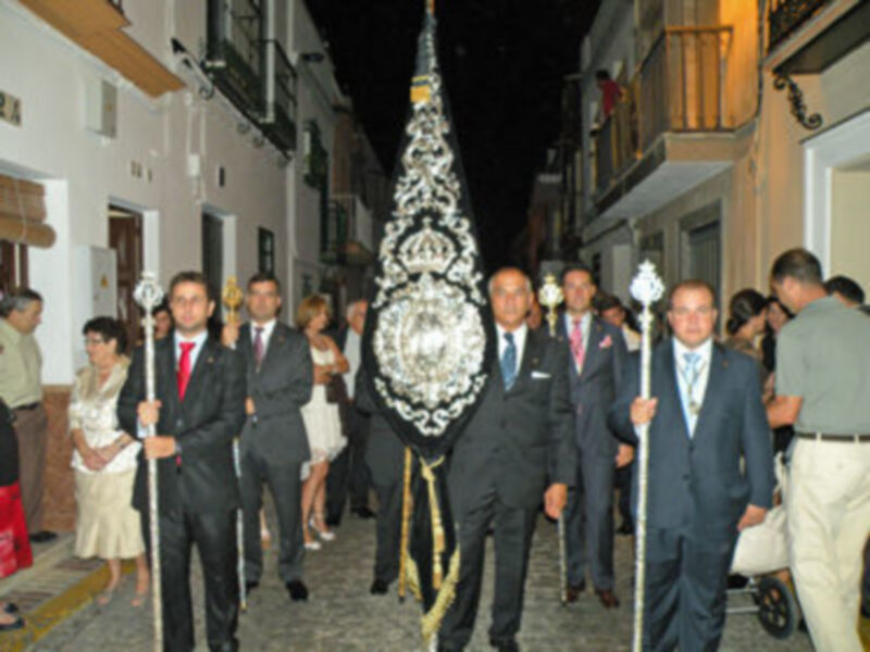 Fiesta religiosa con procesión de portadores de una estandarte ornada en una calle urbana iluminada por la noche.