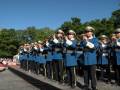 Un grupo de músicos militares en uniformes azules y gorras blancas tocan instrumentos como trompetas, tubas y flautas durante un desfile. El cielo azul claro y árboles verdes forman el fondo de la escena.