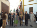 Fotografía de una procesión religiosa con personas vestidas formalmente. En el centro, un grupo de hombres sostiene una bandera con decoraciones doradas y un cetro. La gente observa la procesión, algunas sonriendo o mirando hacia el frente. El entorno muestra un edificio con una puerta y ventana, probablemente de un templo o iglesia.