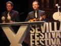 Dos hombres en trajes, uno tocando una guitarra y el otro con instrumentos de percusión, en un escenario con letras que dicen "Sevilla Festival".