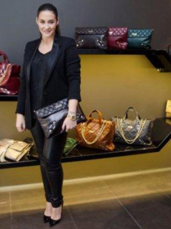 Una mujer con un look elegante de blazer negro, pantalones de piel y zapatos negros posa frente a una vitrina con varios bolsos de lujo.