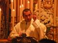 Un sacerdote oficia una ceremonia religiosa con velas y flores en el fondo.