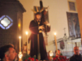 En una iglesia, un hombre en traje negro habla frente a una imagen de Cristo con cruz. A su lado, flores y velas encendidas iluminan la atmósfera.