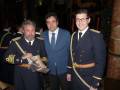 Tres hombres posan juntos en una imagen festiva, dos de ellos con uniformes militares y el tercero en traje formal. La escena parece ser de un evento ceremonial o formal, posiblemente en una sala decorada con luces y adornos.