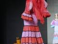 Elegante vestido de flamenca con volantes en tonos rojos y púrpura. La modelo lleva un tocado floral y pendientes largos.