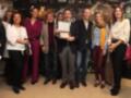 El pié de foto muestra a una persona sosteniendo un premio mientras otros asistentes observan. La escena se desarrolla en un ambiente formal, posiblemente una ceremonia o evento de premiación. La imagen incluye a varias personas vestidas formalmente, con algunos hombres y mujeres de diferentes edades. La iluminación es adecuada para una foto, destacando a los protagonistas y el entorno.