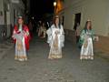 Tres mujeres vestidas con trajes tradicionales caminan por una calle de la noche. La imagen muestra detalles de su vestimenta y el ambiente nocturno de la calle.