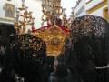 Una procesión religiosa en Andalucía, España. Las personas llevan velos negros y una figura dorada con símbolos religiosos. El cielo es claro, y la arquitectura colonial se ve en el fondo.