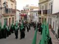 Procesión con nazarenos en túnicas negras y capas verdes, caminando por una calle adoquinada en un entorno urbano.
