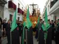 Una procesión religiosa en un ambiente urbano. Personas vestidas con trajes negros y capuchas verdes llevan una imagen de Cristo en un carro procesional. La escena transmite solemnidad y tradición, probablemente durante una celebración religiosa importante.