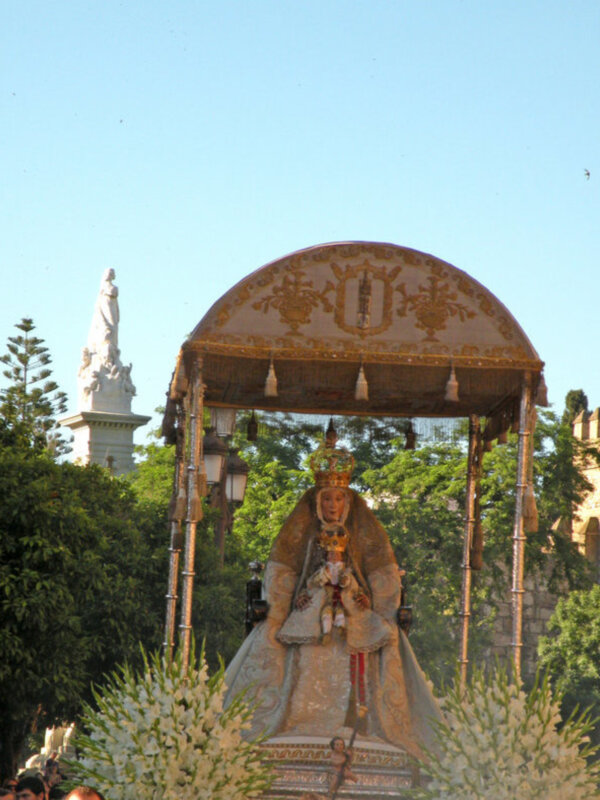 Procesión religiosa con la imagen de la Virgen María en un paso ornado, bajo un cielo azul claro.