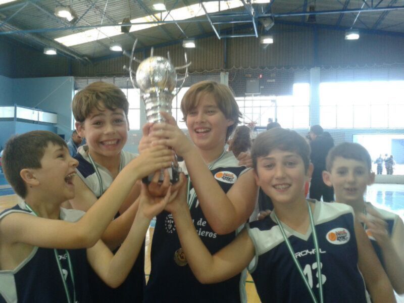 Un grupo de niños sonríen y sostienen una copa dorada en un estadio deportivo. La imagen muestra a los niños con camisetas azules y blancas, uno de ellos sostiene la copa mientras los demás sonríen. La iluminación es brillante y el fondo muestra una estructura de metal en la parte superior.