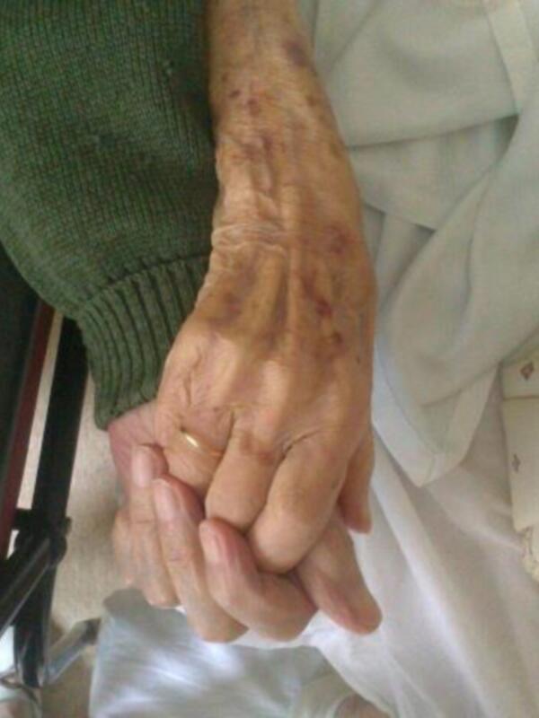 Dos manos se tocan, una de las cuales lleva un anillo. La mano izquierda parece mayor y más vieja, con manchas oscuras en la piel. La mano derecha es más joven y tiene una apariencia más clara. Ambas manos están en una posición que sugiere un gesto de apoyo o amistad.