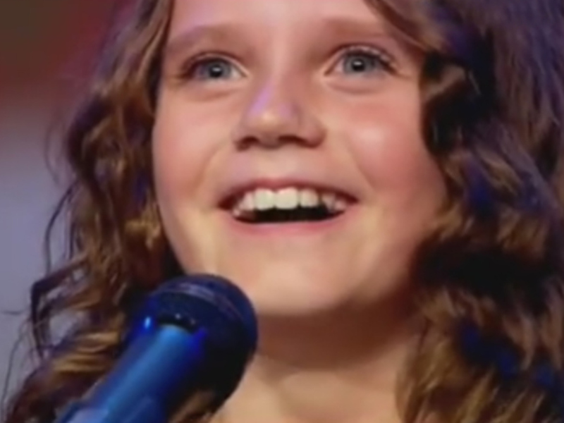 Una niña con cabello rizado y una sonrisa amplia, frente a un micrófono.