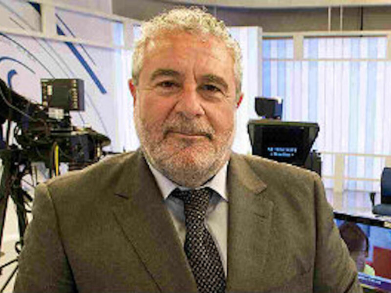 Un hombre en un entorno profesional, posiblemente una sala de televisión o estudio de grabación. La imagen muestra a un hombre con cabello blanco y barba, vestido con una chaqueta de color claro. En el fondo se pueden ver elementos típicos de un estudio de televisión, como cámaras y pantallas.