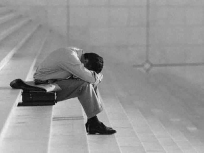 Un hombre con la cabeza entre las manos, sentado en una escalera de hormigón. La escena es sombría y minimalista, enfocándose en la expresión de desesperanza o tristeza del individuo.