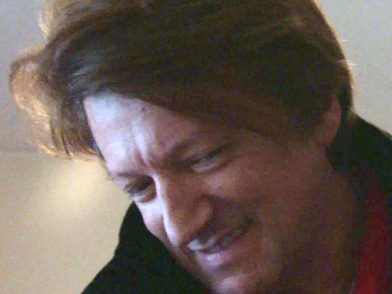 Una imagen de una persona con cabello largo y ondulado, mirando hacia abajo. La persona lleva una chaqueta de color verde oscuro y un cuello rojo. El fondo es neutro, probablemente una pared blanca o de color claro.