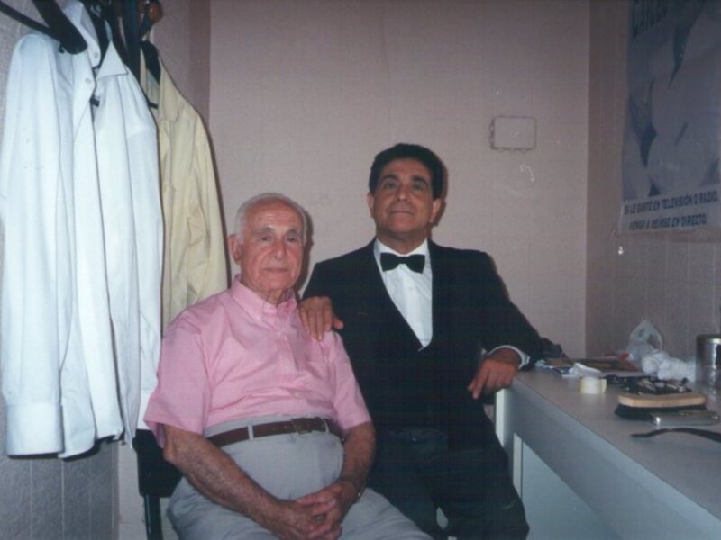 Dos hombres posan juntos en una habitación con camisas colgadas en el fondo.