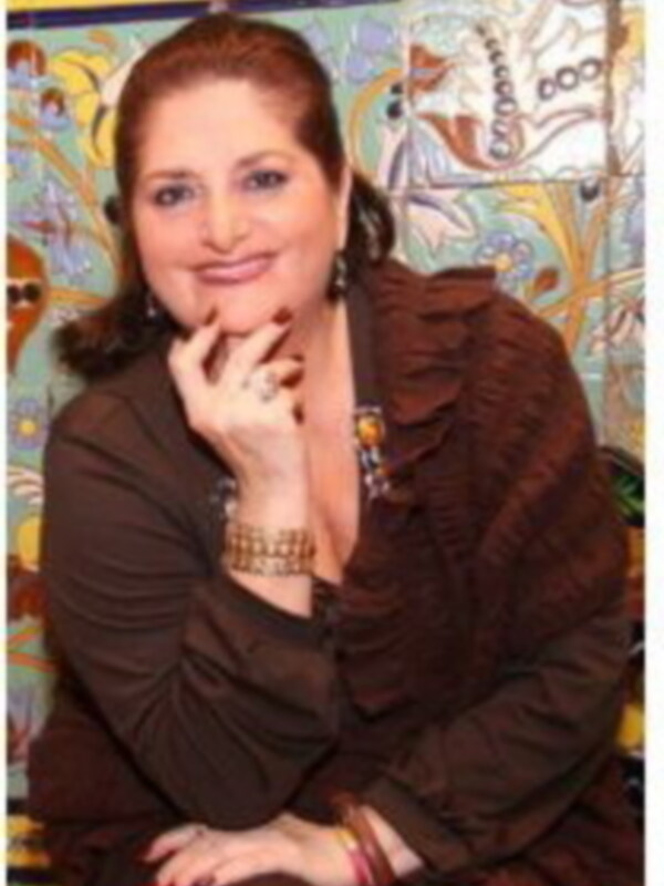 Una mujer con una blusa marrón y un collar de perlas, posando con las manos en la cadera.