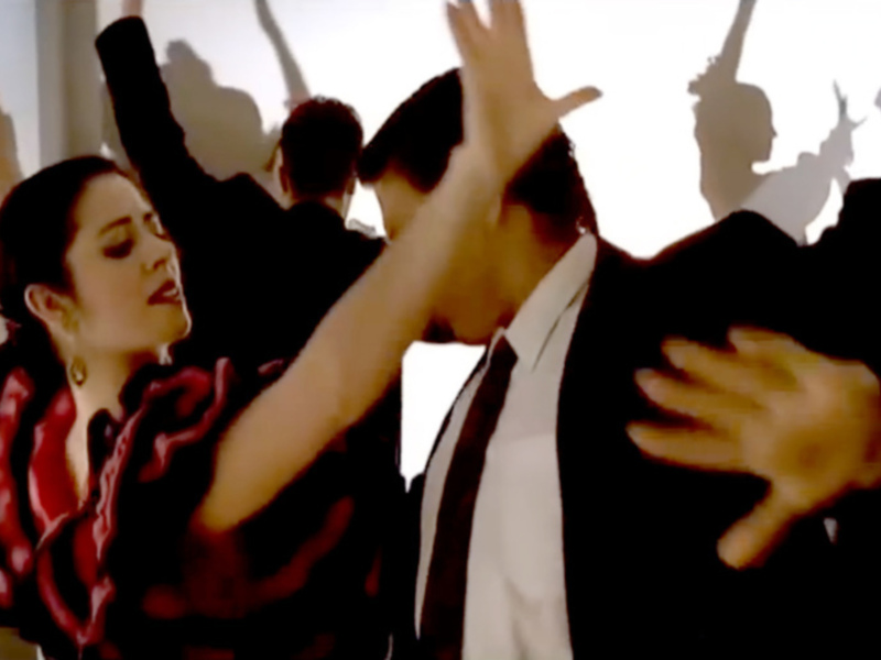 Una pareja baila con entusiasmo en un ambiente brillante y desenfadado. La imagen captura el movimiento vibrante de sus manos y la expresión de alegría en su rostro.