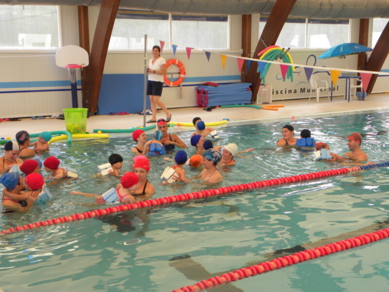 Niños aprendiendo a nadar en una piscina interior. El agua es clara y los niños están usando gorros de natación. Hay banderas colores en el fondo, una canasta y un baloncesto cerca de la pared.