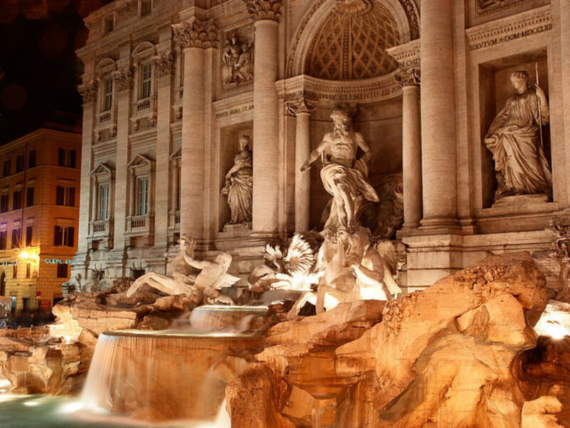 Fotografía nocturna de la famosa fuente de Trevi en Roma, Italia. La imagen muestra una escultura de un dios acuático con un pez en la mano, flanqueada por dos figuras femeninas. La fuente está rodeada de rocas y cascadas, iluminada por la luz de las farolas. La arquitectura clásica y los detalles escultóricos son visibles en el entorno.