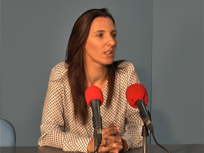 A una mujer con cabello largo y ondulado, vestida con un traje de chaqueta a cuadros blanco y negro. Se encuentra en una sala con fondo azul, frente a micrófonos rojos y negros. La mujer parece estar en una entrevista o conferencia de prensa, con expresión seria y gesto serio.
