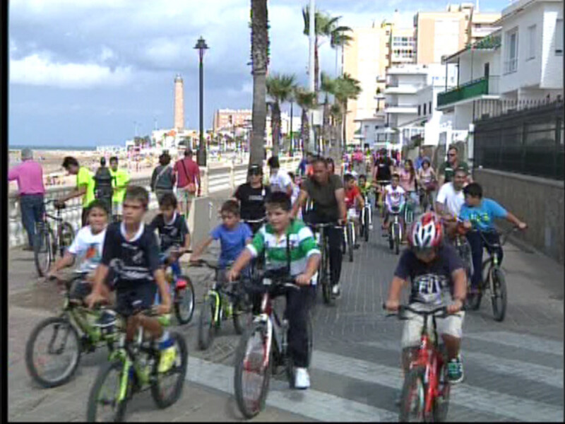 Un grupo de niños y adultos en bicicletas disfrutan de un día soleado por la playa, con edificios y palmeras al fondo.