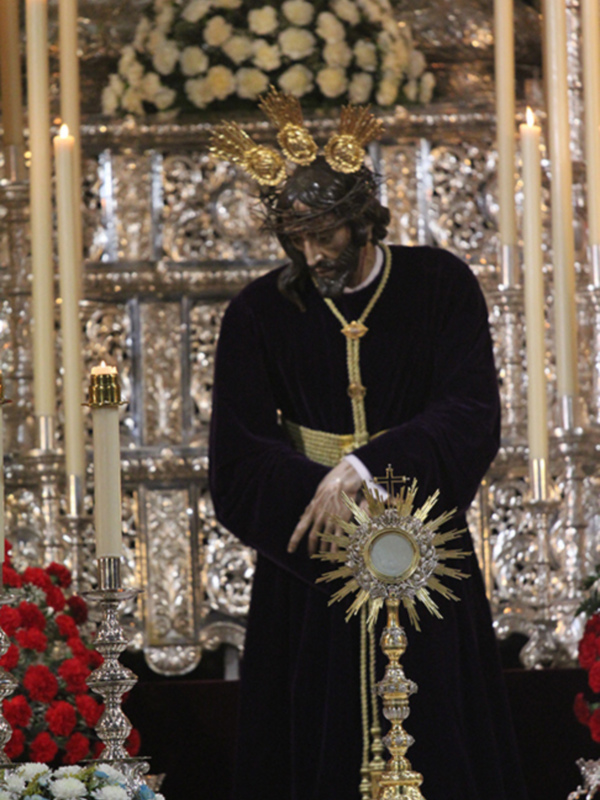 Procesión religiosa con imagen de Jesús Nazareno en trono dorado, rodeado de flores y velas.