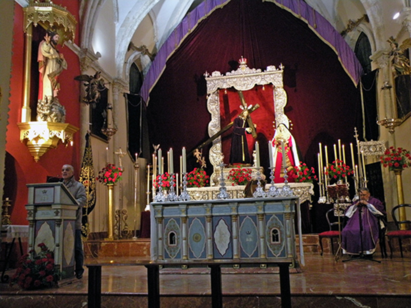 Interior de una iglesia con altares adornados, presbiterio y sacerdotes en actitud de oración.
