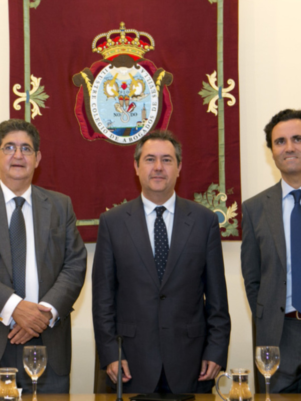 Tres hombres en trajes formales posan en una sala con un escudo de armas en el fondo.